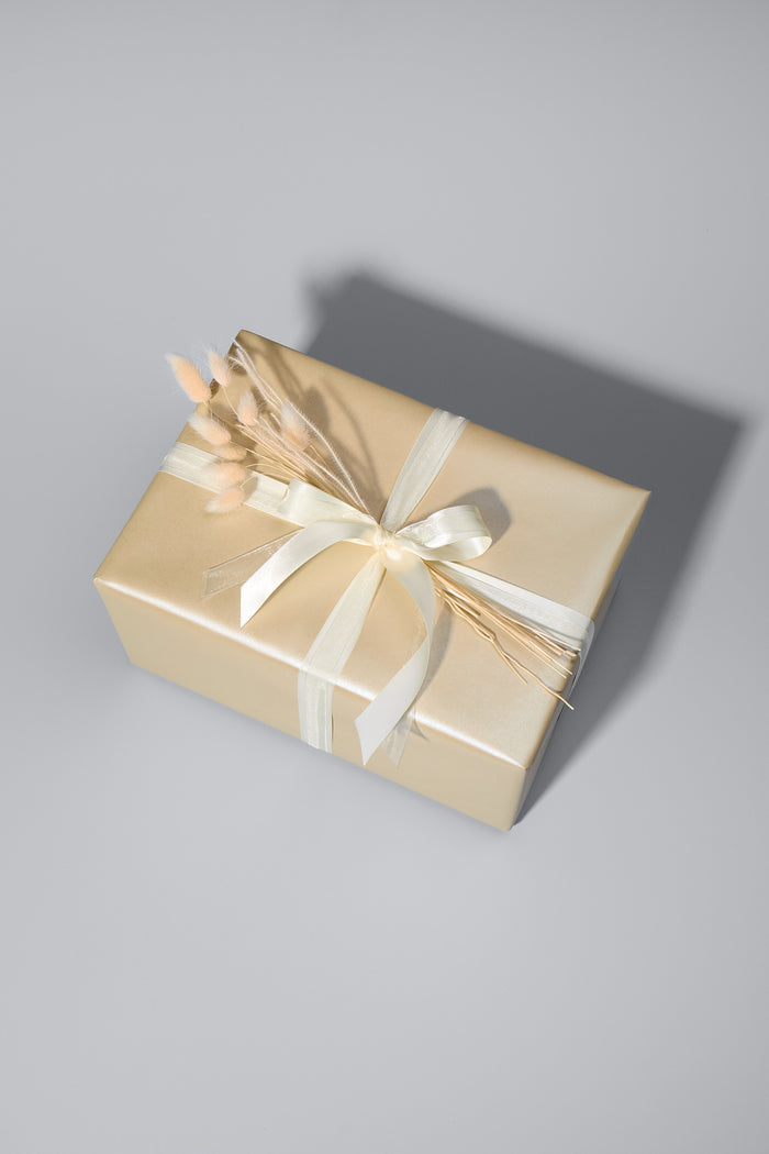 Ein stilvoll verpacktes Geschenk in beigem Papier mit edlem Schimmer. Es ist mit zwei cremefarbenen Schleifen gebunden und mit zarten Trockenblumen verziert. Das Geschenk liegt auf einer hellgrauen Oberfläche, während sein Schatten für eine elegante Tiefe sorgt.