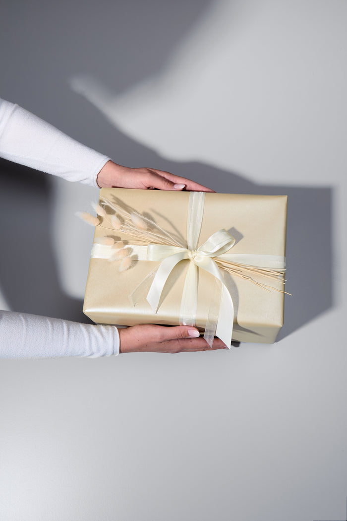 Ein stilvoll verpacktes Geschenk in edlem, schimmerndem beigem Papier mit zwei cremefarbenen Schleifen und zarten Trockenblumen als Dekoration. Zwei Hände in einem weissen Langarmshirt halten das Geschenk, als würde es überreicht werden. Der helle Untergrund und die weichen Schatten verleihen dem Bild eine elegante Atmosphäre.