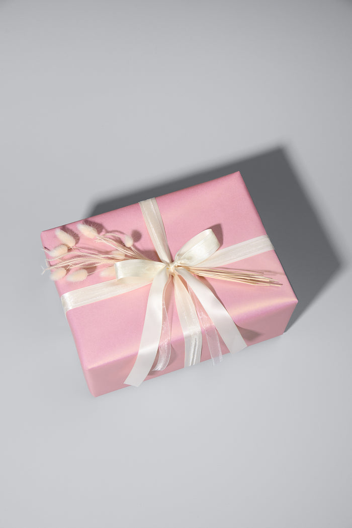 Ein stilvoll verpacktes Geschenk in rosafarbenem Papier mit schimmernder Optik. Es ist mit zwei cremefarbenen Schleifen gebunden und mit zarten Trockenblumen dekoriert. Das Geschenk liegt auf einer hellgrauen Oberfläche, während sein Schatten eine elegante Tiefe verleiht.