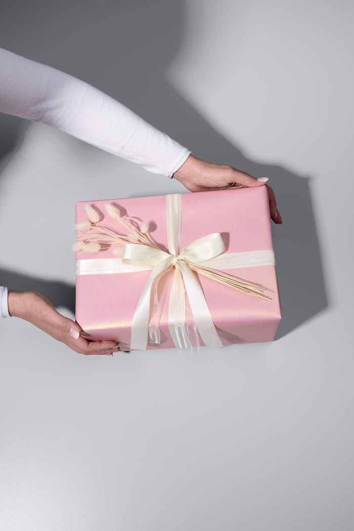 Ein stilvoll verpacktes Geschenk in edlem, schimmerndem rosafarbenem Papier mit zwei cremefarbenen Schleifen und zarten Trockenblumen als Dekoration. Zwei Hände in einem weissen Langarmshirt halten das Geschenk, als würde es überreicht werden. Der helle Untergrund und die weichen Schatten verleihen dem Bild eine elegante Atmosphäre.