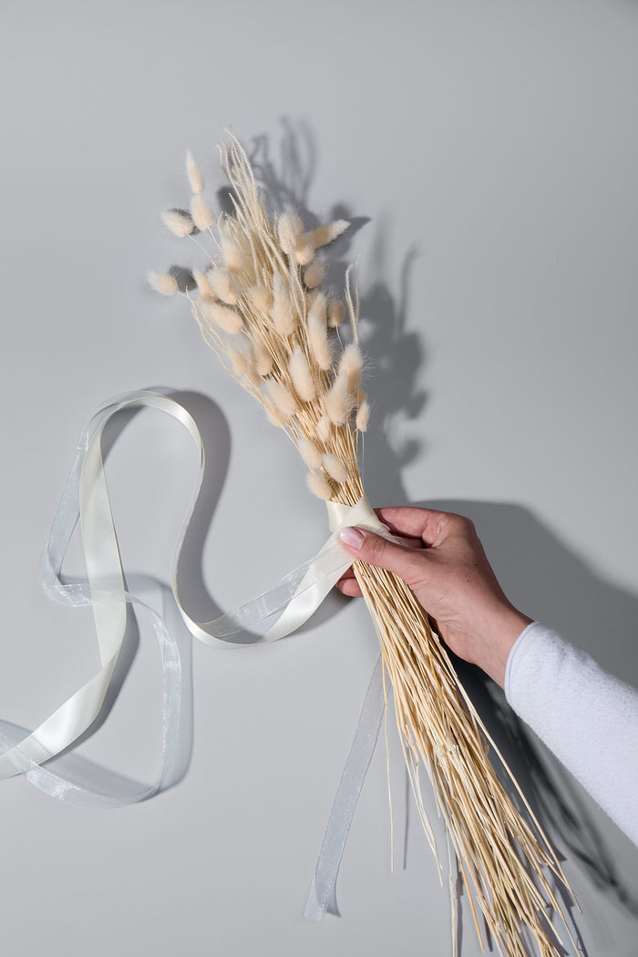 Eine Hand in einem weissen Langarmshirt hält einen Bund zarter, cremefarbener Trockenblumen, der mit zwei Schleifen – einer aus glänzendem Satin und einer transparenten – gebunden ist. Die Bänder fallen elegant über die hellgraue Oberfläche und verstärken die edle, minimalistische Ästhetik des Bildes.