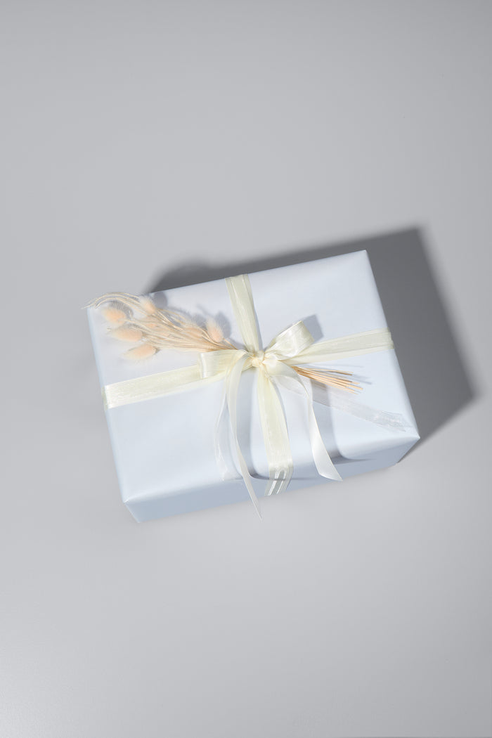 Ein stilvoll verpacktes Geschenk in weissem Papier mit edlem Schimmer. Es ist mit zwei cremefarbenen Schleifen gebunden und mit zarten Trockenblumen verziert. Das Geschenk liegt auf einer hellgrauen Oberfläche, während sein Schatten für eine elegante Tiefe sorgt.