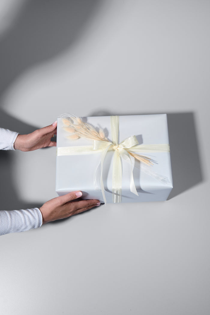 Ein stilvoll verpacktes Geschenk in edlem, schimmerndem weissem Papier mit zwei cremefarbenen Schleifen und zarten Trockenblumen als Dekoration. Zwei Hände in einem weissen Langarmshirt halten das Geschenk, als würde es überreicht werden. Der helle Untergrund und die weichen Schatten verleihen dem Bild eine elegante Atmosphäre.